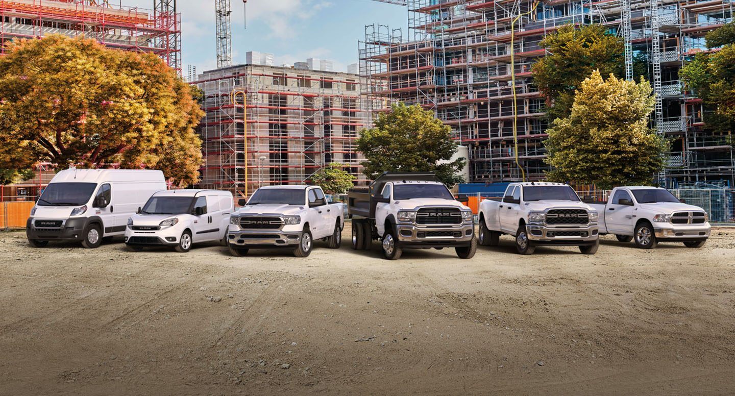 A lineup of six 2022 Ram trucks and vans in front of a tall building under construction, covered in scaffolding.
