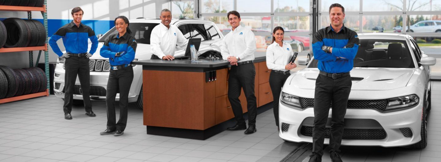 The Service department in a dealership with six employees standing near the service desk with two white vehicles--one on each side of the desk.