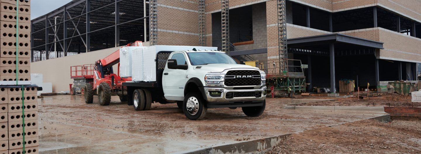 A white 2024 Ram 5500 SLT Chassis Cab Regular Cab with a platform upfit loaded with building materials, parked at a commercial construction site.